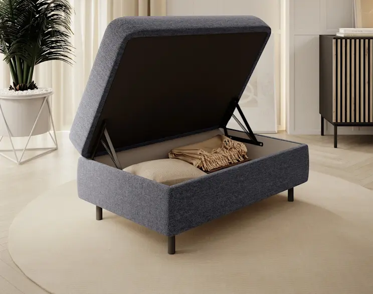 Otwierana pufa Gomsi z pojemnikiem obok szafki RTV Meorati, ustawiona w jasnym, eleganckim salonie z naturalnymi dodatkami i miękkim dywanem.