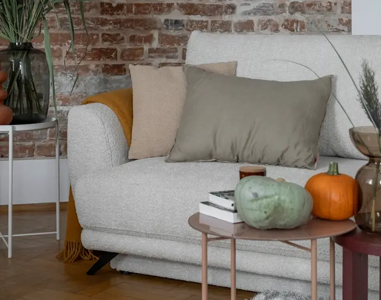 Kanapa Megis w jasnej tkaninie boucle, ozdobiona poduszkami i kocem, w jesiennej aranżacji z dyniami i dekoracjami na stolikach pomocniczych.