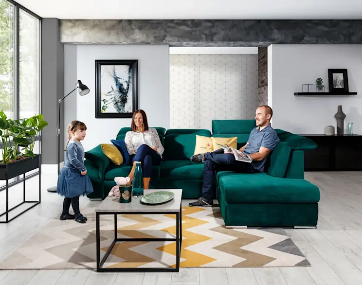 Rodzinny salon z narożnikiem Loreto Kronos 19 w zielonej tkaninie, jasnym dywanem w geometryczny wzór i rodziną spędzającą czas razem w przytulnym wnętrzu