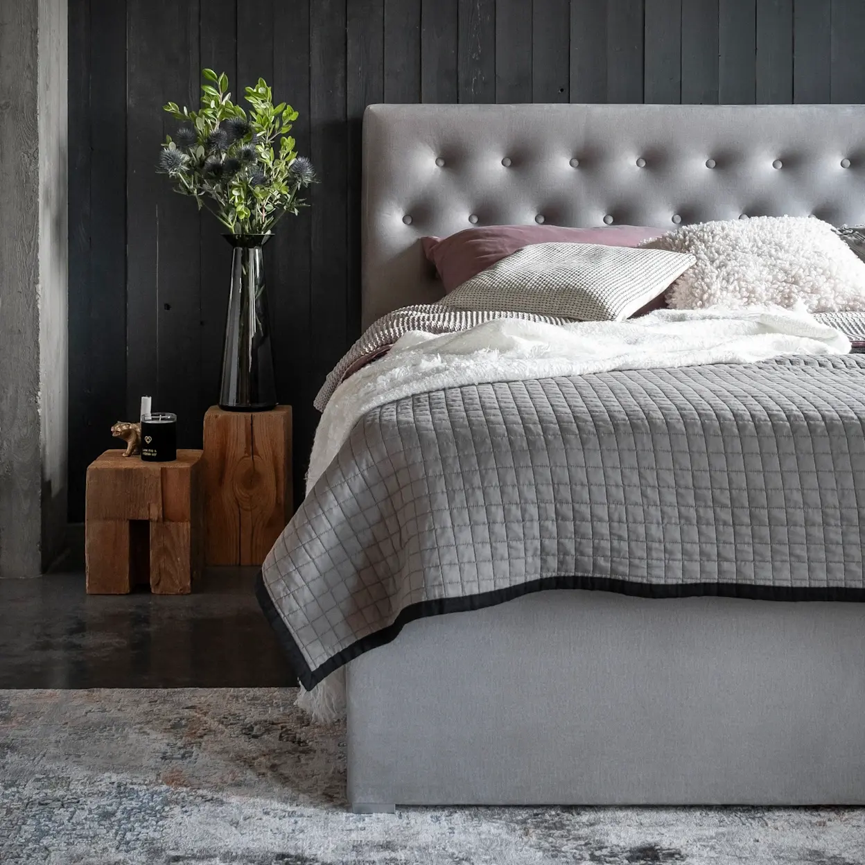 Łóżko kontynentalne Loree z pojemnikiem w nowoczesnej sypialni. Pikowane wezgłowie, szara tapicerka i ciemna ściana tworzą elegancki klimat.