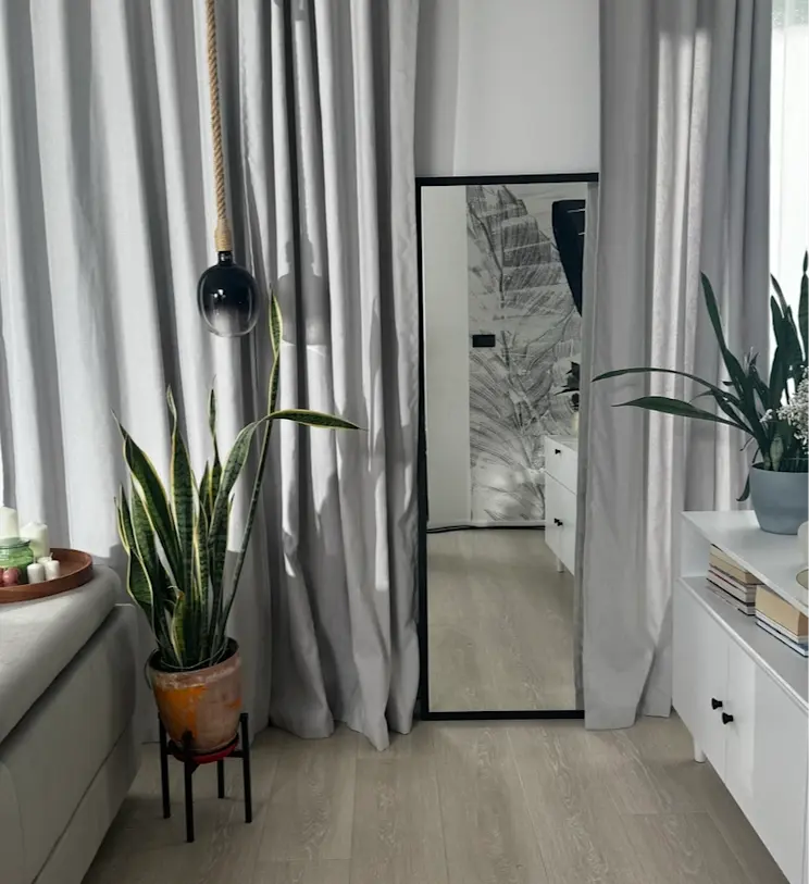 Lustro Aruba w czarnej ramie ustawione przy ścianie z szarymi zasłonami, obok roślin w donicach, białych mebli Dullum i wiszącej lampy z liną.