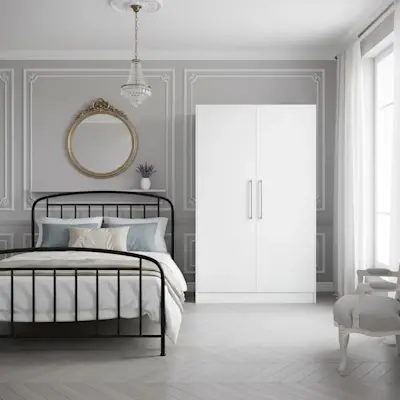 Biała szafa Alarica II w eleganckiej sypialni z klasycznymi zdobieniami na ścianach, metalowym łóżkiem i dekoracyjnym lustrem. Jasna kolorystyka i subtelne detale tworzą stylowe, harmonijne wnętrze sprzyjające relaksowi.