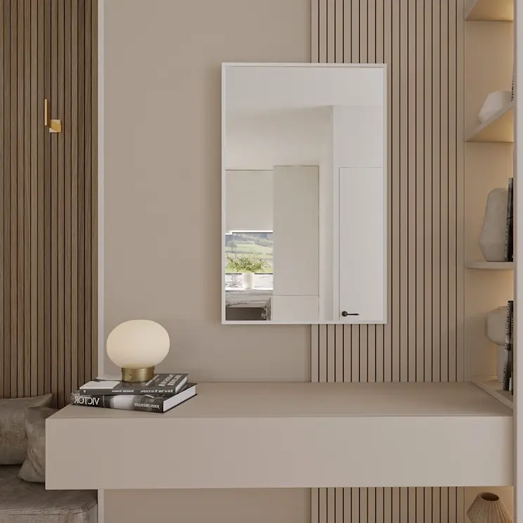 Lustro ścienne Fellan prostokątne w białej ramie zawieszone na tle pionowych lameli. Pod lustrem znajduje się minimalistyczna toaletka, a na blacie leży stos książek i stoi kulista lampka.