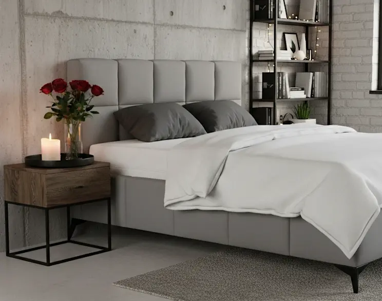 Tapicerowane łóżko Mist Nube 03 w szarym kolorze w nowoczesnej sypialni w stylu loft. Obok stoi drewniana szafka nocna z metalową podstawą, na której znajduje się świeca i czerwone róże w wazonie.