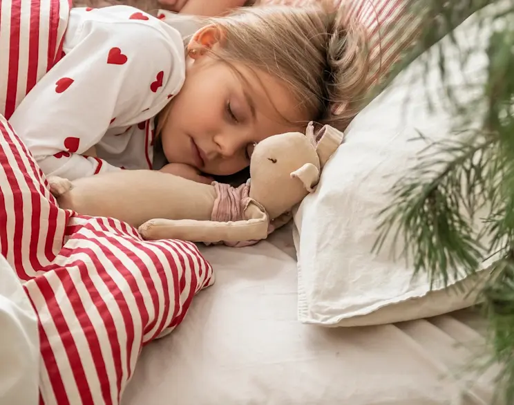 Dziecko śpi w łóżku Dizzle, przytulone do pluszowej zabawki, w jasnej pościeli. Kadr pokazuje świąteczny, domowy klimat i komfortowy wypoczynek w sypialni, idealny dla rodziny z dziećmi.