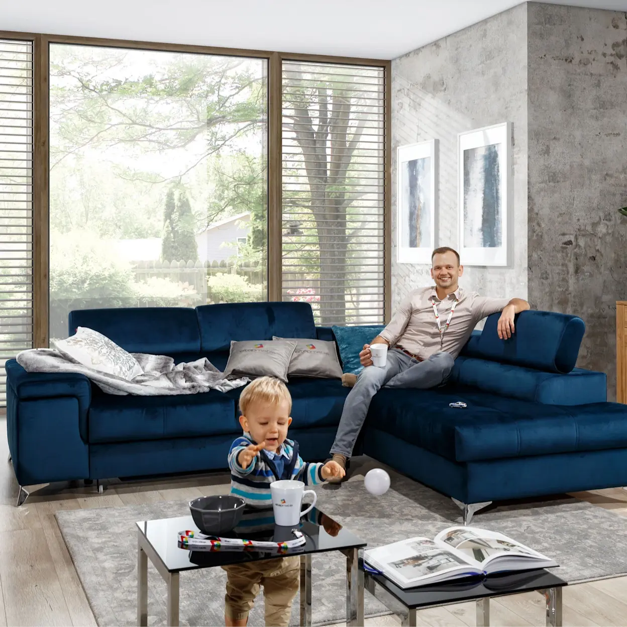 Granatowy narożnik Eridano Monolith 77 z funkcją spania w nowoczesnym salonie; miękkie poduszki, duże przeszklenia i szara ściana w stylu loft tworzą przytulną przestrzeń dla rodziny.