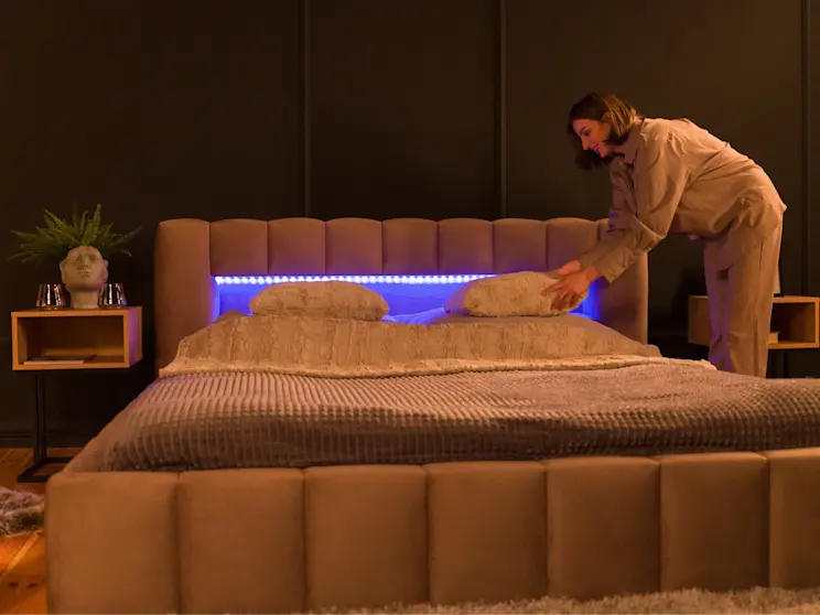 Tapicerowane łóżko Lamica w tkaninie Monolith 09 z podświetleniem LED w zagłówku, w ciemnej sypialni. Kobieta układa poduszki na beżowej pościeli, a obok stoją stoliki nocne z dekoracjami.