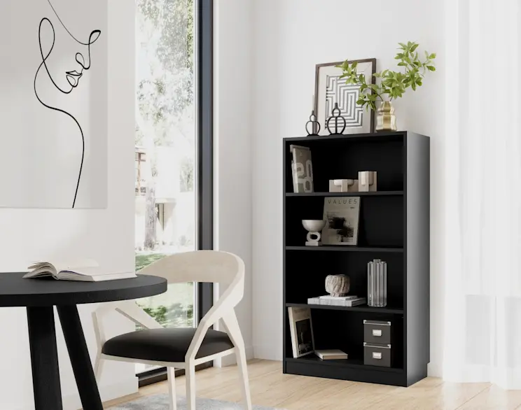 Czarny regał Pocett z otwartymi półkami w jasnym wnętrzu przy dużym oknie. Na półkach książki i dekoracje, na górze wazon z zieloną gałązką i grafika, obok okrągły stół i krzesło.