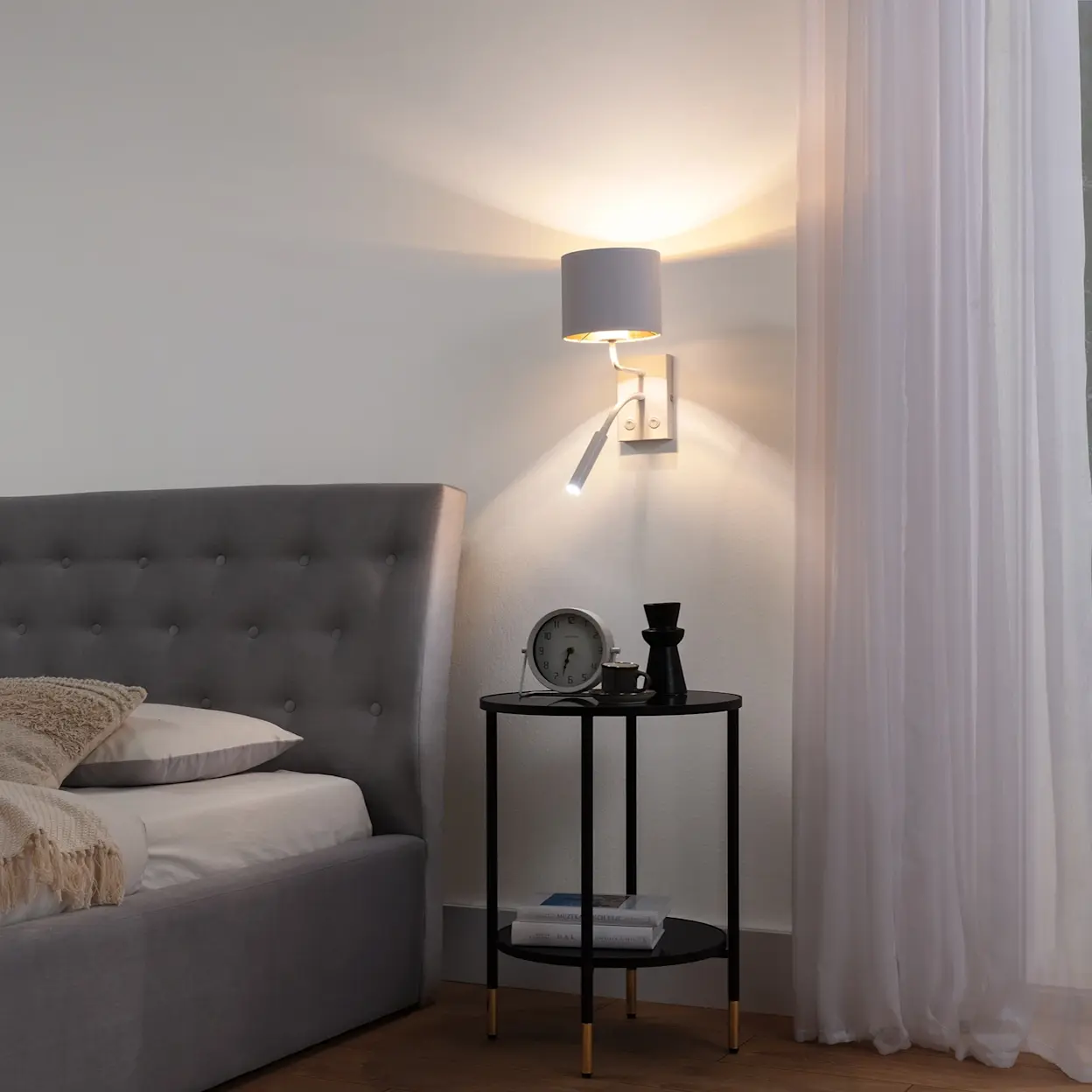 Sypialnia z tapicerowanym łóżkiem Milano w szarości i kinkietem Vespillo nad stolikiem nocnym, miękkie światło przy zasłonach.