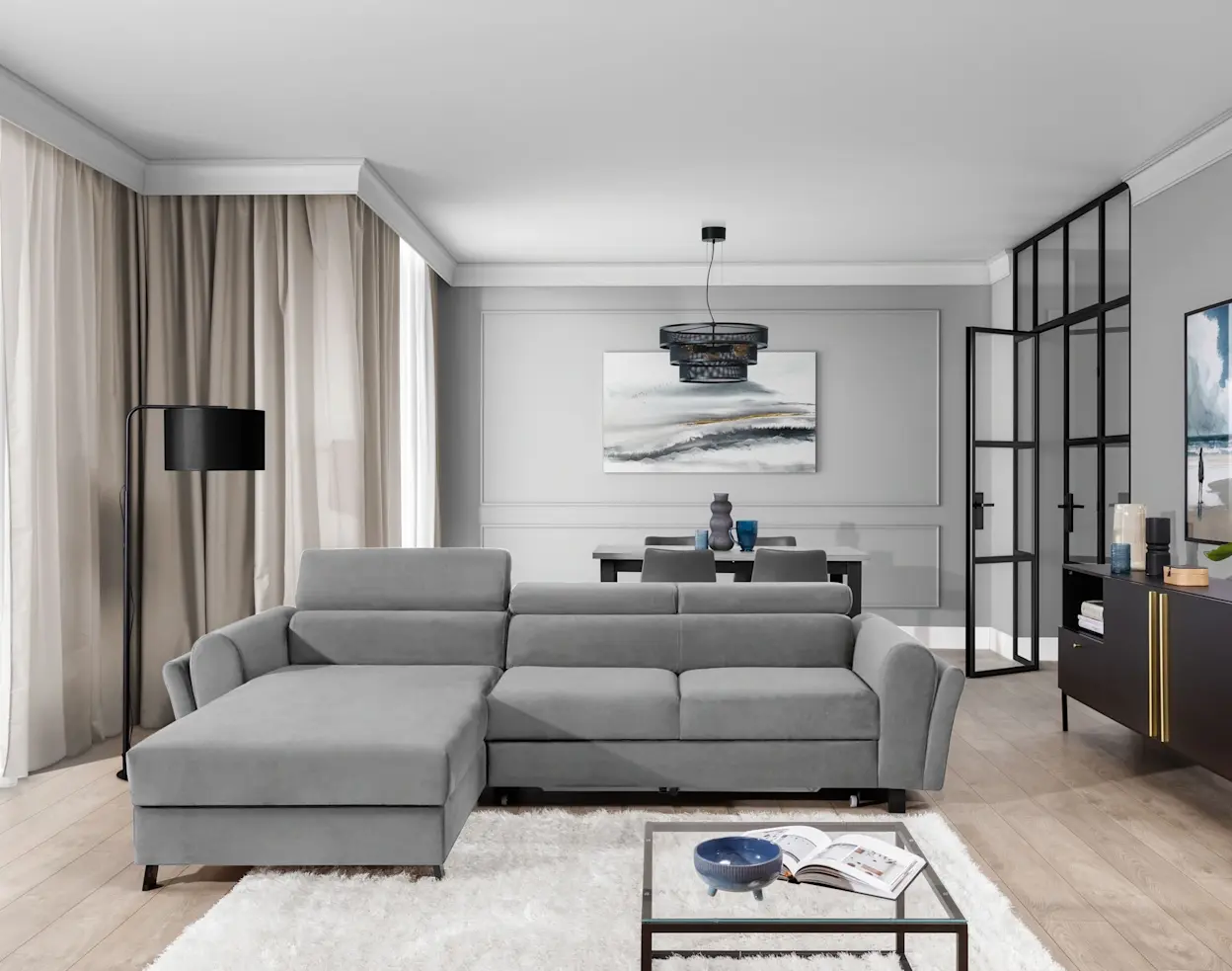 Przykład aranżacji salonu z szarym narożnikiem Mariall i szezlongą po lewej stronie, pomocny przy planowaniu wymiarów. W tle widać zasłony, lampę podłogową, stół z krzesłami oraz przeszklone drzwi.