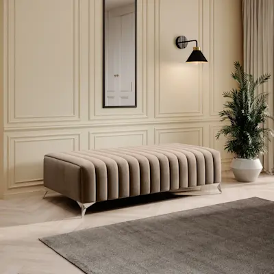 Beżowa pufa Laurence w tkaninie Monolith 76 ustawiona w eleganckim wnętrzu z jasną sztukaterią. Nad nią wisi lustro Arizona w czarnej ramie, obok świeci kinkiet Detmer, a na podłodze leży dywan Verlice Grey w stonowanym odcieniu.