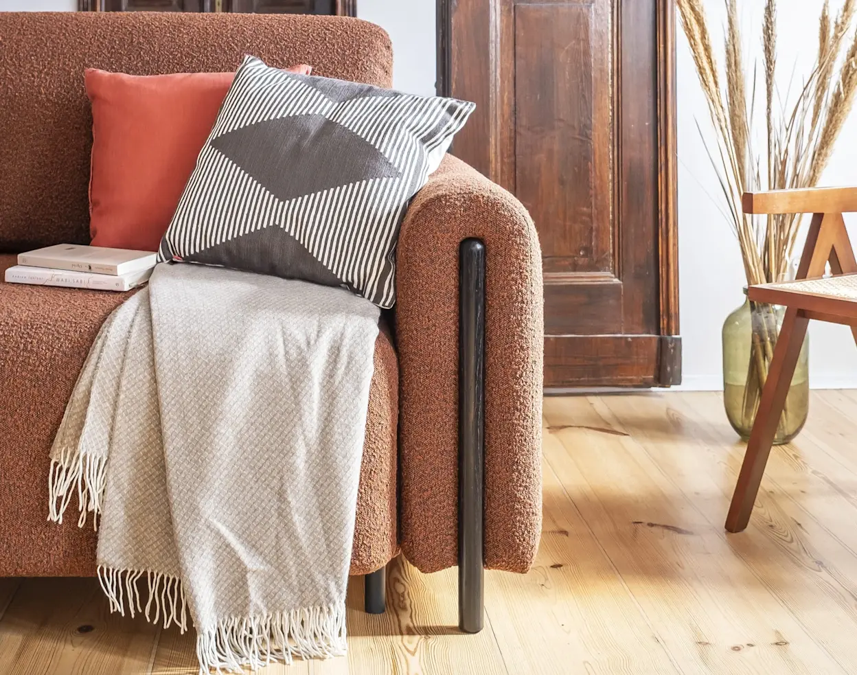 Zbliżenie na kanapę Arcadova Jaffray 30 w ciepłym odcieniu, ozdobioną poduszkami i pledem, w przytulnym wnętrzu z drewnianymi akcentami.