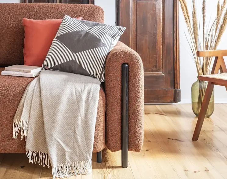 Zbliżenie na kanapę Arcadova Jaffray 30 w ciepłym odcieniu, ozdobioną poduszkami i pledem, w przytulnym wnętrzu z drewnianymi akcentami.