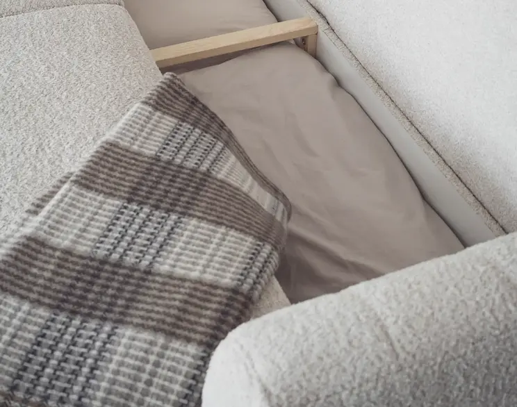 Zbliżenie na funkcję spania w kanapie Megis Royal 18: rozłożone siedzisko z widoczną powierzchnią do spania i drewnianym elementem konstrukcji. W kadrze widać jasną tkaninę boucle oraz koc w kratę w neutralnych kolorach.