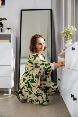 Uśmiechnięta kobieta w kwiecistym stroju porządkuje rzeczy w białej komodzie, przy dużym lustrze i dekoracjach w przytulnej sypialni.