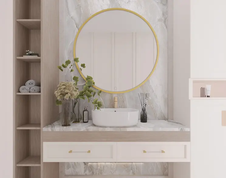 Aranżacja łazienki z okrągłym lustrem Gerbinie w złotej ramie zawieszonym nad białą umywalką nablatową. Wnętrze uzupełnia jasna szafka z uchwytami w kolorze złota, kamienny blat oraz dekoracje: roślina, wazon i dyfuzor zapachowy.
