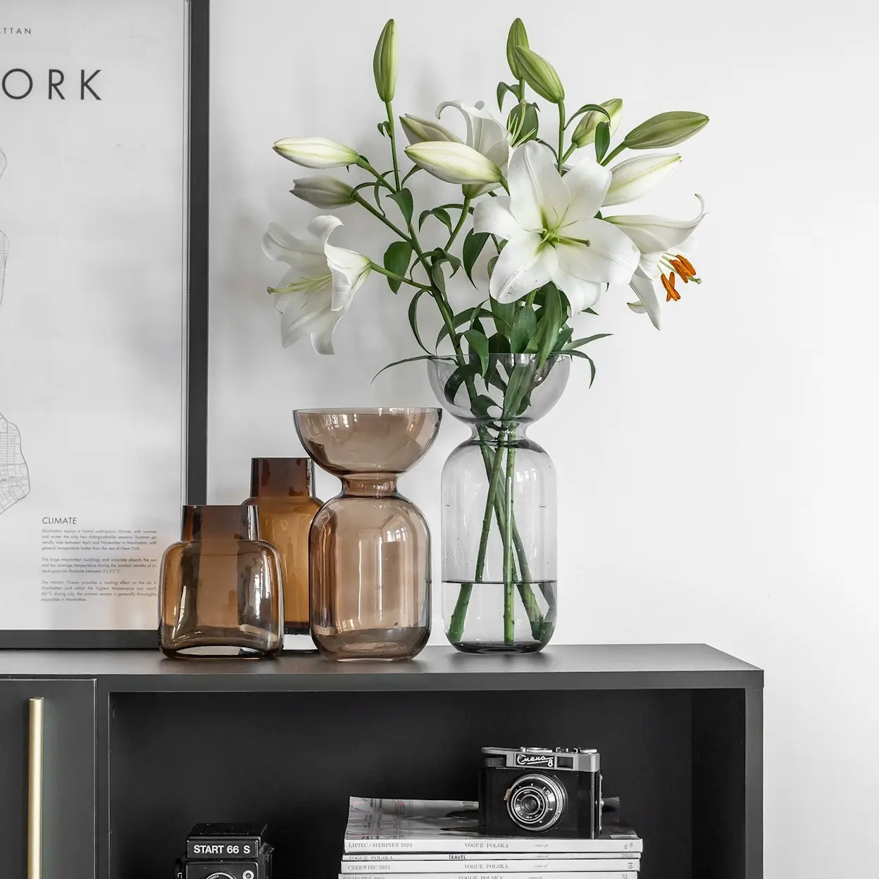 Zbliżenie na czarną komodę Lanzzi ze złotą listwą i otwartą półką. Na blacie stoją szklane wazony w odcieniach bursztynu oraz wazon z białymi liliami, a w półce widać aparat i stos magazynów. W tle jasna ściana z plakatem w czarnej ramie.