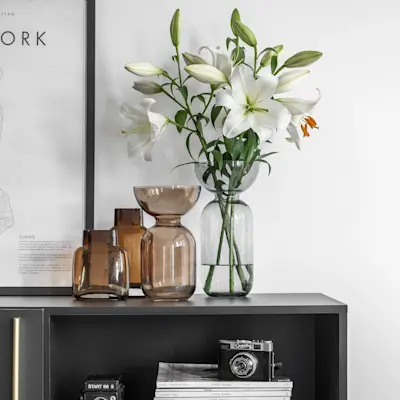 Zbliżenie na czarną komodę Lanzzi ze złotą listwą i otwartą półką. Na blacie stoją szklane wazony w odcieniach bursztynu oraz wazon z białymi liliami, a w półce widać aparat i stos magazynów. W tle jasna ściana z plakatem w czarnej ramie.