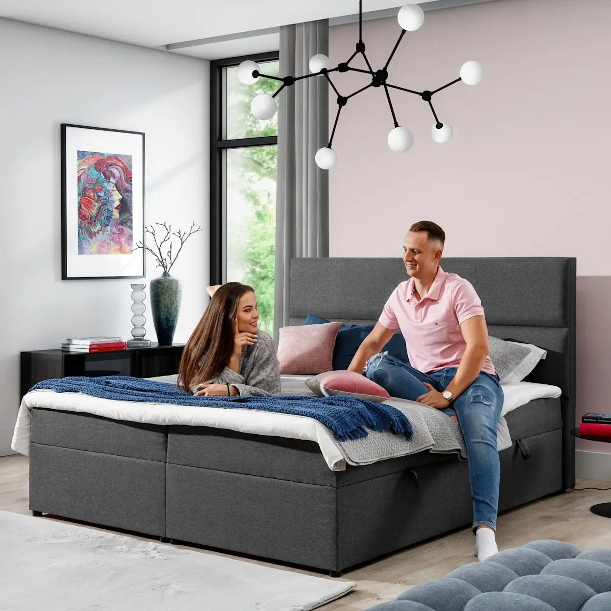 Szare łóżko kontynentalne Amber 140x200 w tkaninie Sawana 05 w nowoczesnej sypialni. Na łóżku siedzi para, widać wysoki zagłówek, poduszki i koc, a nad nim designerskie oświetlenie.