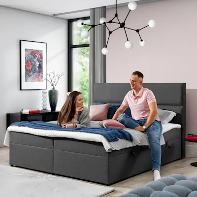 Szare łóżko kontynentalne Amber 140x200 w tkaninie Sawana 05 w nowoczesnej sypialni. Na łóżku siedzi para, widać wysoki zagłówek, poduszki i koc, a nad nim designerskie oświetlenie.