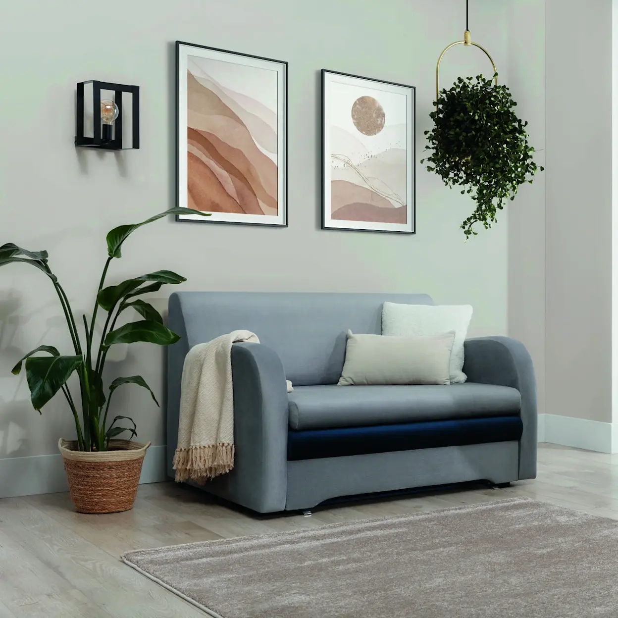 Nowoczesne wnętrze z kanapą Ario Nube 03/Monolith 76 ustawioną na dywanie w jasnym salonie. Ujęcie pokazuje kinkiet Giorri na ścianie, wiszącą lampę Giorri z rośliną oraz dekoracyjne grafiki, które dodają przytulnego klimatu.