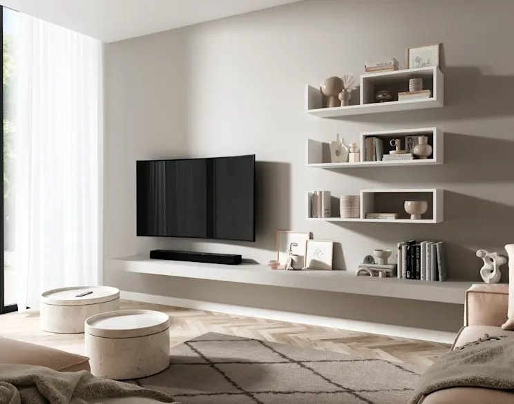 Biała półka ścienna Vellia w nowoczesnym salonie, obok strefy TV i długiej zabudowy, z książkami i dekoracjami w jasnych barwach.