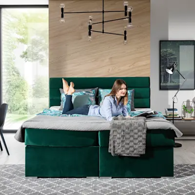 Zielone łóżko kontynentalne Softy z pojemnikiem w tkaninie Monolith 37 w nowoczesnej sypialni. Na łóżku leży narzuta i poduszki, a całość uzupełnia drewniana ściana, designerska lampa sufitowa i przytulny dywan.