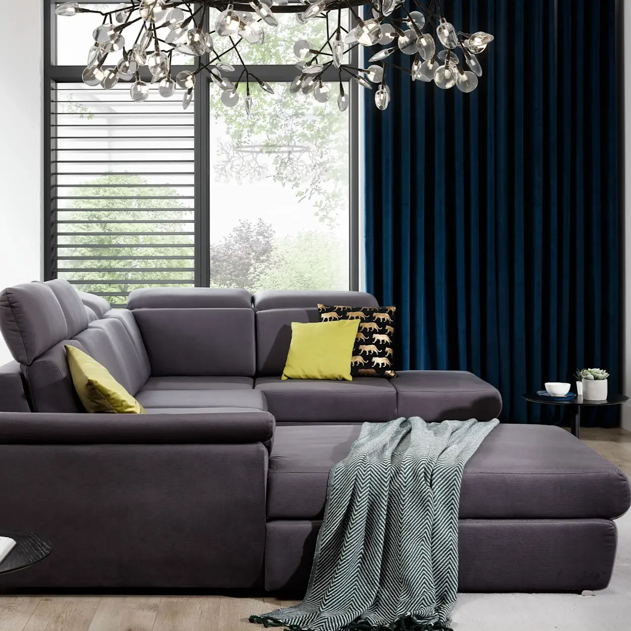 Nowoczesny narożnik Evanell z funkcją spania w eleganckim salonie. Grafitowa tapicerka, dekoracyjne poduszki i stylowe oświetlenie.