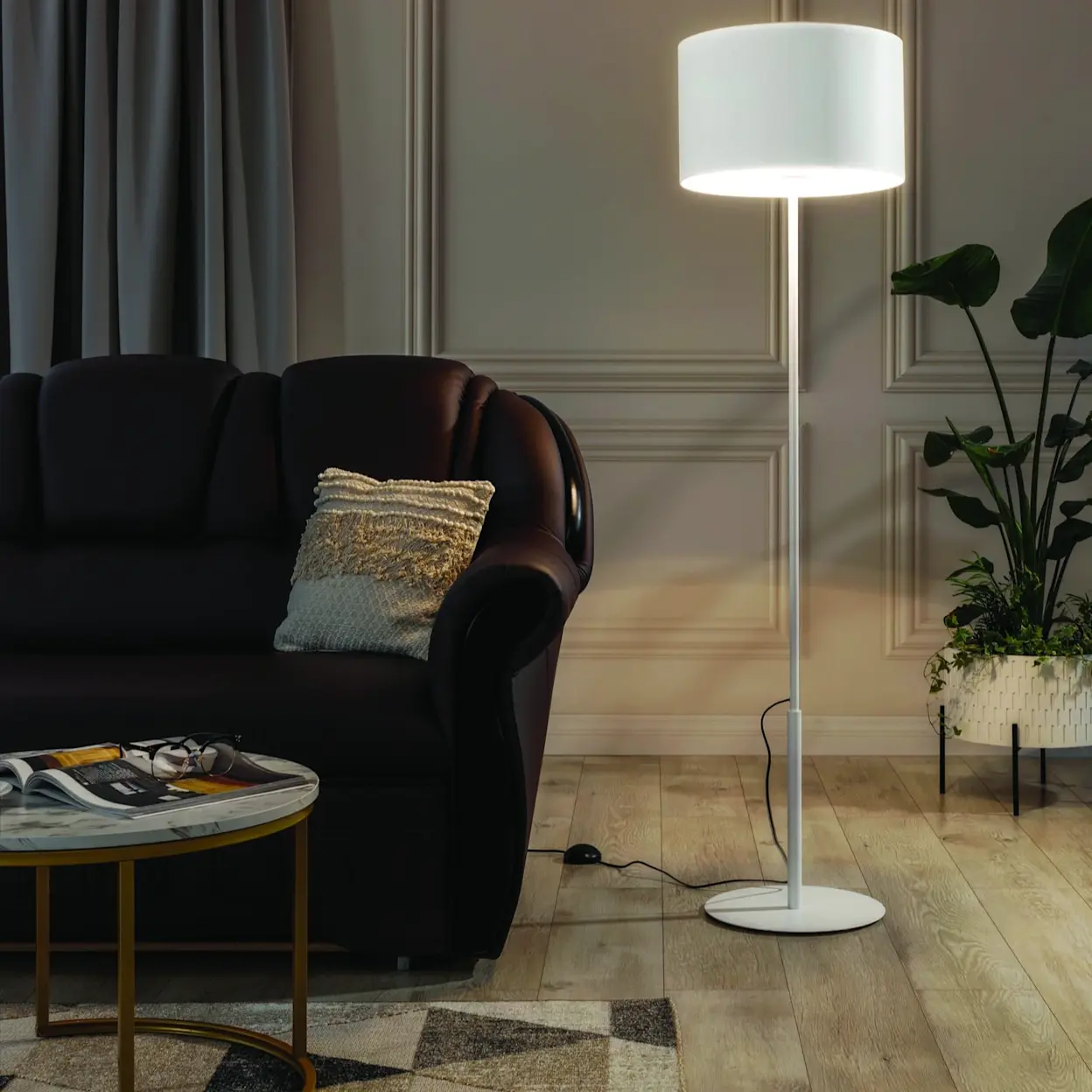 Biała lampa stojąca Vespillo z dużym abażurem doświetla strefę wypoczynku w salonie. Obok znajduje się narożnik Lord w ciemnej tapicerce, a na podłodze leży dywan Eustache Beige oraz stolik z okrągłym blatem z czasopismem.