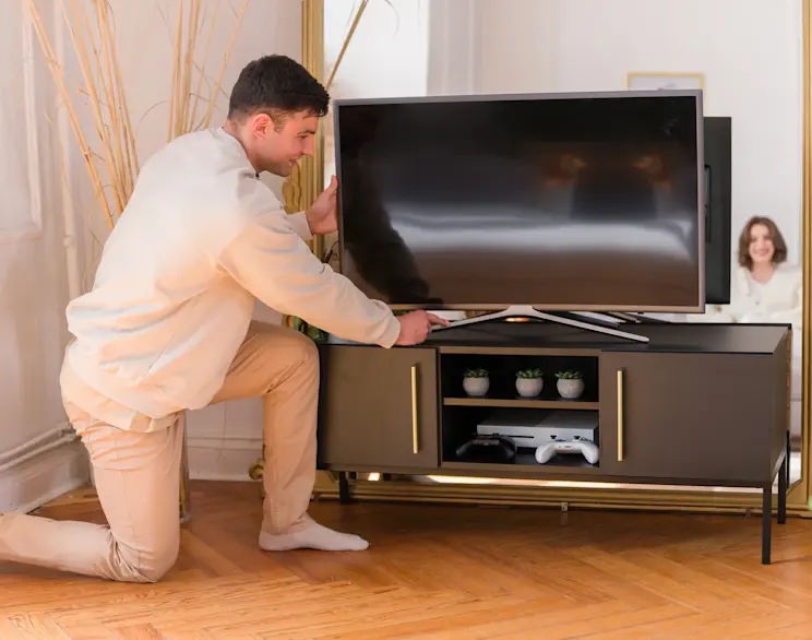 Czarna szafka RTV Lanzzi ze złotymi uchwytami, na której ustawiany jest telewizor, w jasnym wnętrzu z drewnianą podłogą i dużym lustrem.