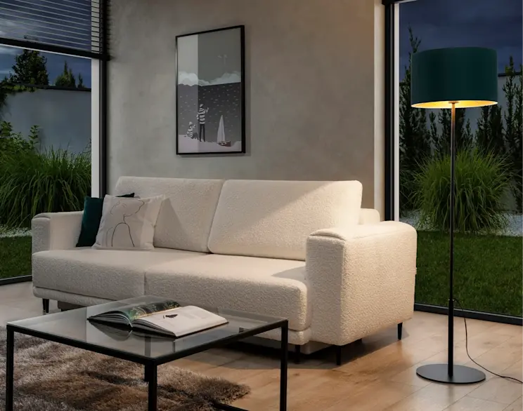 Lampa stojąca Vespillo z zielonym abażurem i czarną podstawą ustawiona przy jasnej sofie w tkaninie boucle. W kadrze widać też szklany stolik kawowy, dywan oraz duże okna z widokiem na ogród, a ciepłe światło lampy buduje przytulny nastrój.