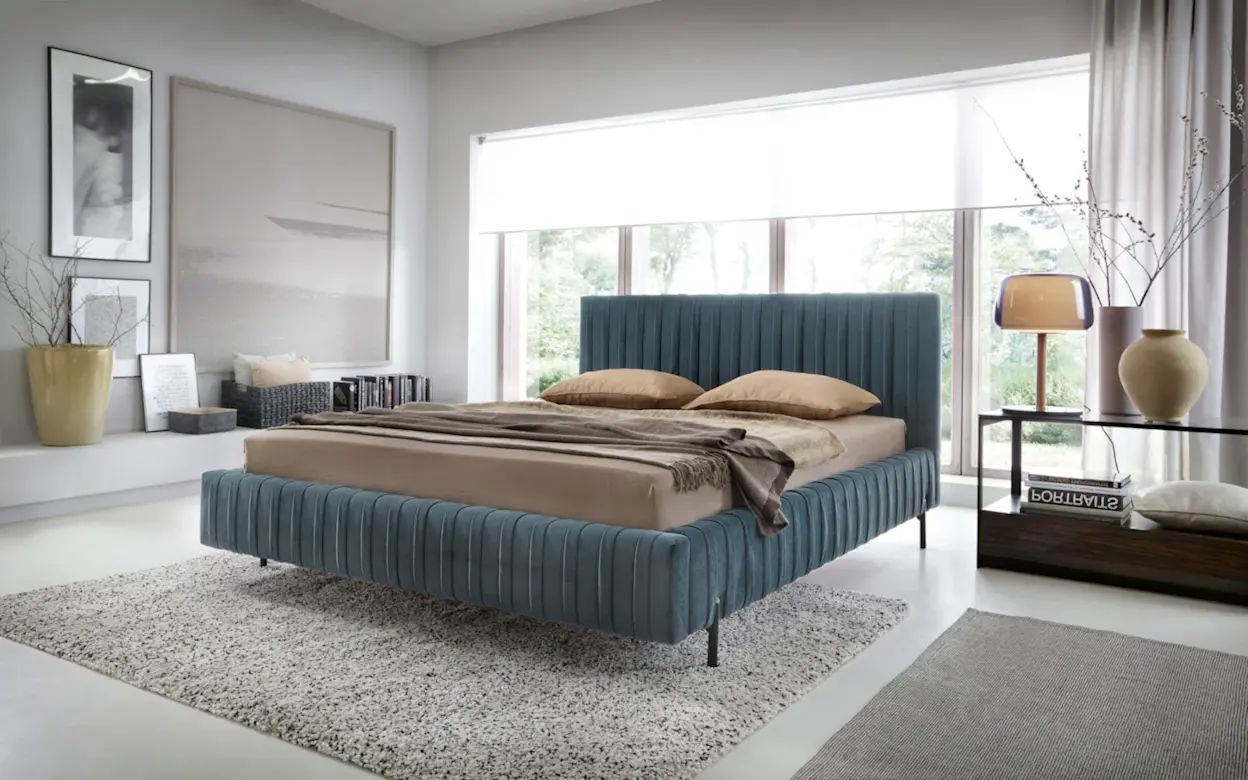 Łóżko Plissa Velvetmat 100 w nowoczesnej, jasnej sypialni z dużymi oknami, miękkim dywanem i eleganckimi dodatkami w naturalnych barwach.