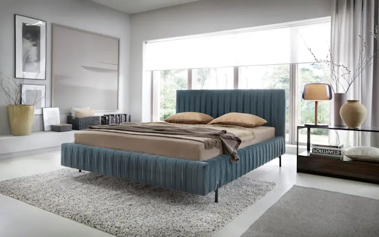 Łóżko Plissa Velvetmat 100 w nowoczesnej, jasnej sypialni z dużymi oknami, miękkim dywanem i eleganckimi dodatkami w naturalnych barwach.