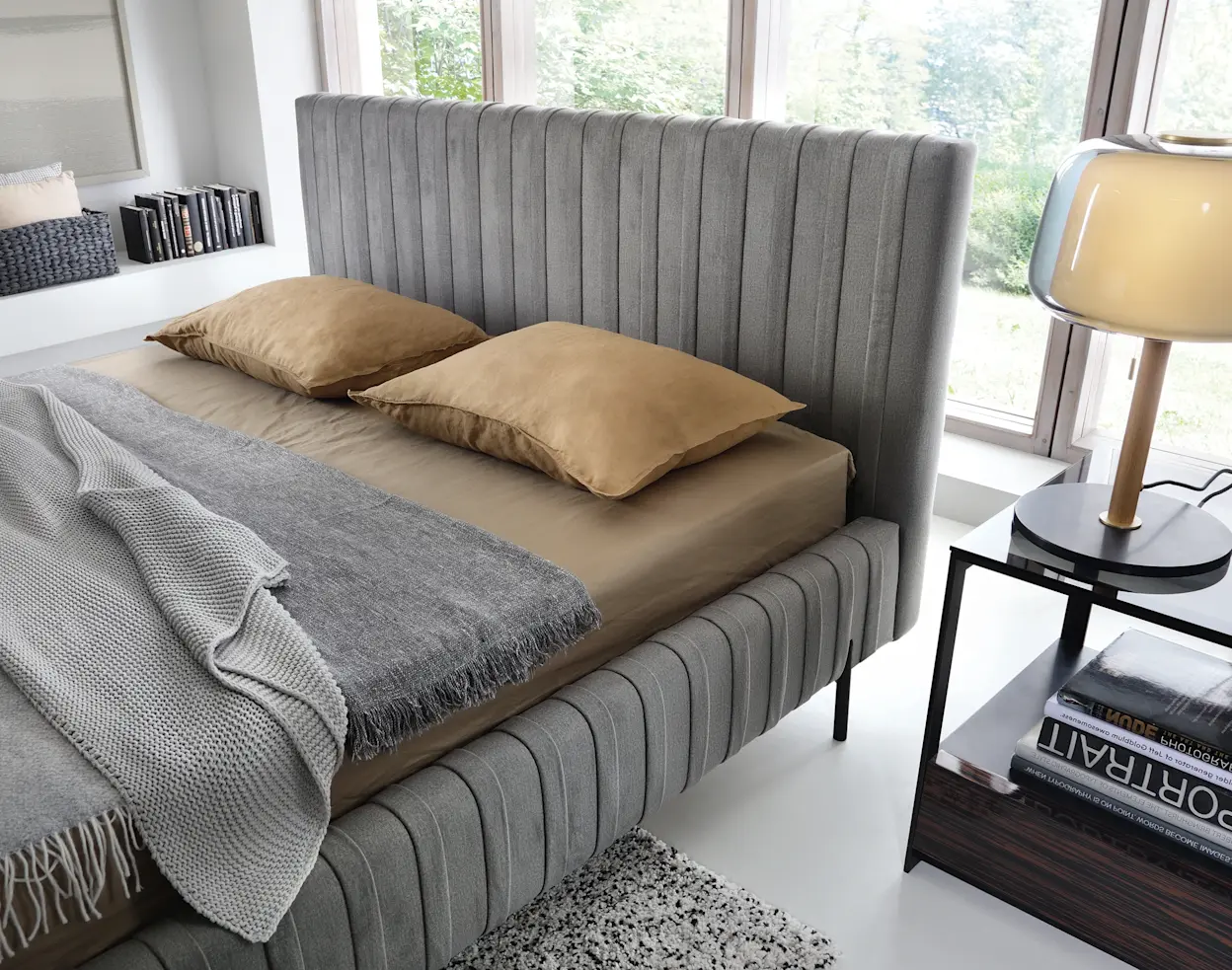 Łóżko Plissa z wysokim, panelowym wezgłowiem i tapicerowaną ramą, ustawione w jasnej sypialni z dużym oknem i minimalistycznymi dodatkami.