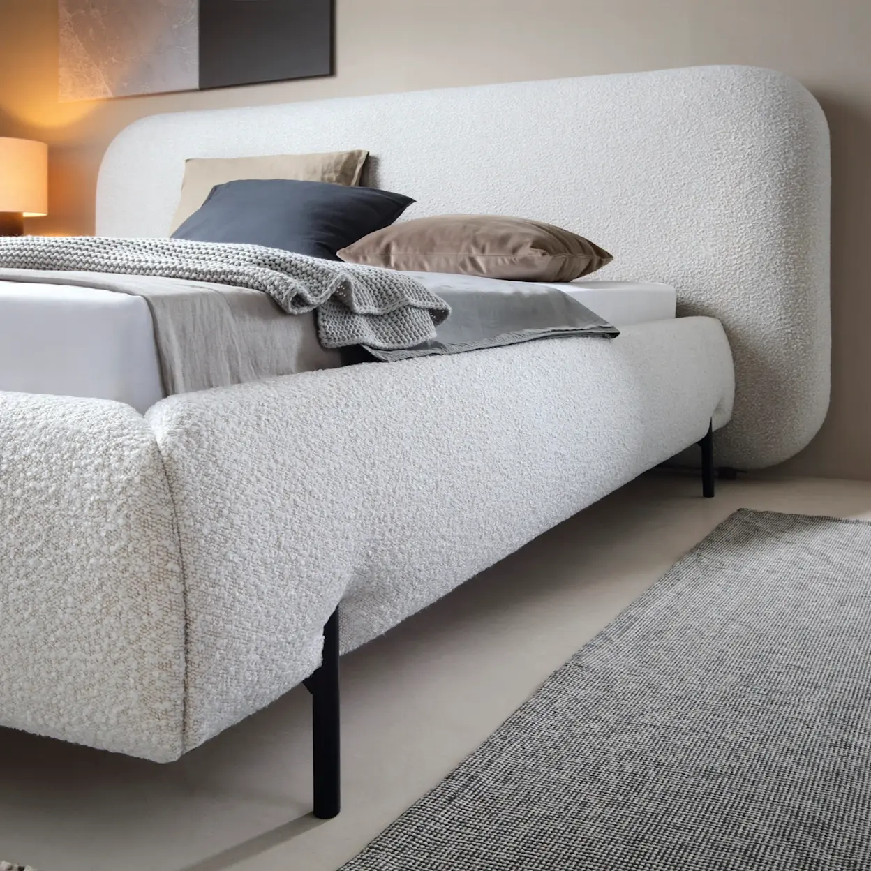 Tapicerowane łóżko Pointt Jaffray 18 w jasnej tkaninie bouclé z dużym, zaokrąglonym zagłówkiem. Na pościeli leżą dekoracyjne poduszki i pled, a czarne, smukłe nóżki dodają lekkości aranżacji sypialni.