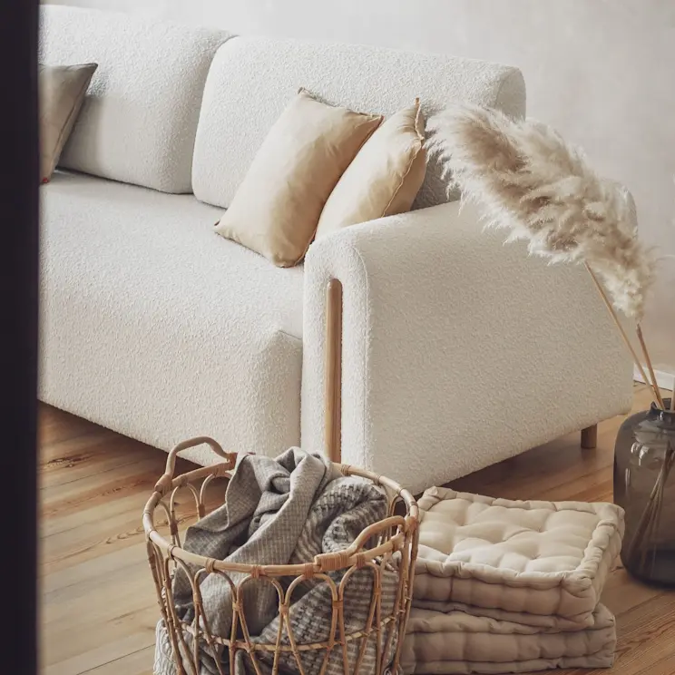 Beżowa kanapa Arcadova z tkaniny boucle, z jasnymi poduszkami i naturalnymi dodatkami, tworząca ciepłą, przytulną aranżację salonu.