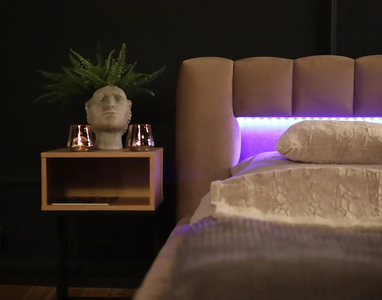 Łóżko Lamica w tkaninie Monolith 09 w przytulnej sypialni z ciemną ścianą. Wysoki, tapicerowany zagłówek ma podświetlenie LED tworzące nastrojową poświatę, a obok stoi stolik nocny z dekoracjami i rośliną.