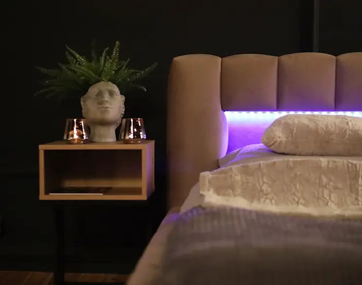 Łóżko Lamica w tkaninie Monolith 09 w przytulnej sypialni z ciemną ścianą. Wysoki, tapicerowany zagłówek ma podświetlenie LED tworzące nastrojową poświatę, a obok stoi stolik nocny z dekoracjami i rośliną.