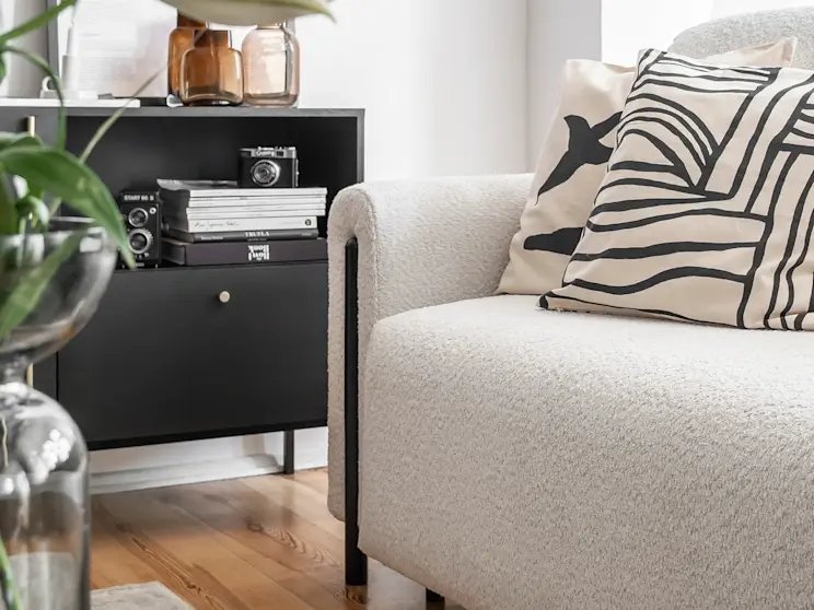 Aranżacja kącika wypoczynkowego z kanapą Arcadova II w jasnej, miękkiej tapicerce oraz czarną komodą Lanzzi. Na komodzie widać stos albumów i aparat, a w tle znajdują się szklane wazony i roślina, tworząc nowoczesny klimat.