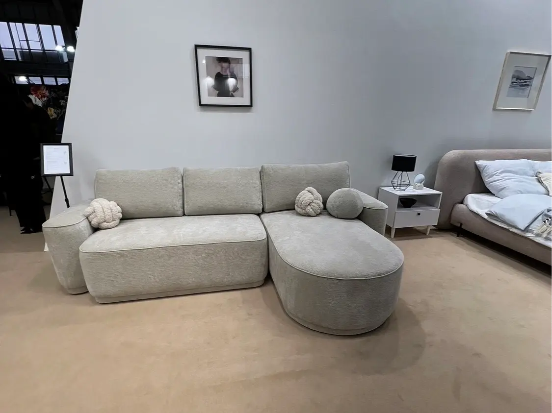 Jasny narożnik z funkcją spania o zaokrąglonej bryle w nowoczesnym wnętrzu. Miękka tapicerka, szerokie siedzisko i wygodna otomana tworzą komfortową strefę relaksu w salonie.