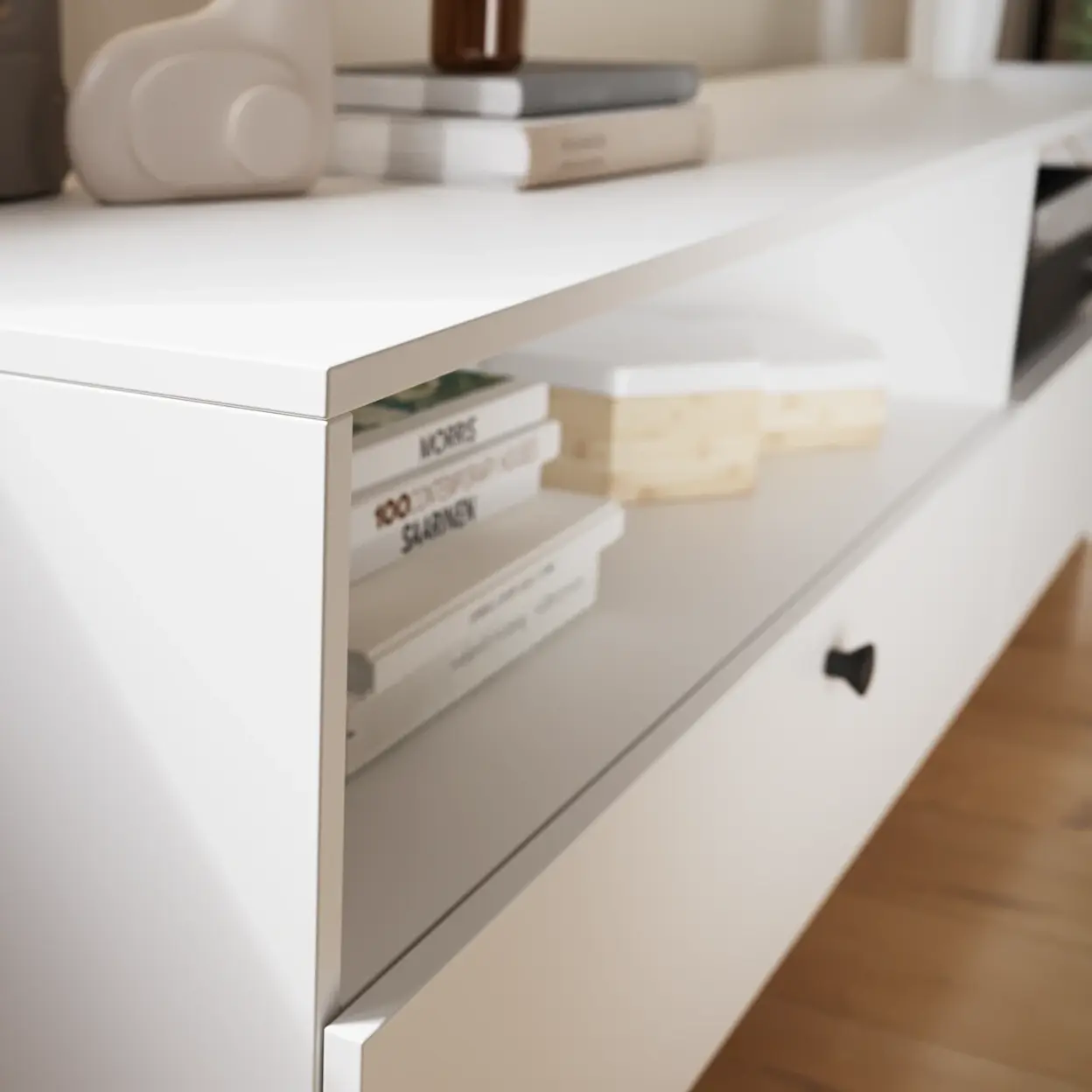 Zbliżenie na białą szafkę RTV Dullum – widoczny gładki blat, otwarta półka z ułożonymi książkami oraz front szuflady z czarnym uchwytem. Minimalistyczny mebel w jasnym wnętrzu, sfotografowany pod kątem, z rozmytym tłem.