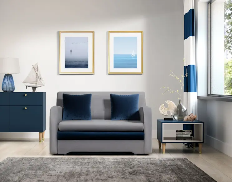 Kanapa Ario w szarym kolorze z granatowymi poduszkami w jasnym salonie w stylu marynistycznym. Wnętrze uzupełniają granatowe meble, dekoracje z motywem żaglówki i jasna podłoga, tworząc spójną aranżację.