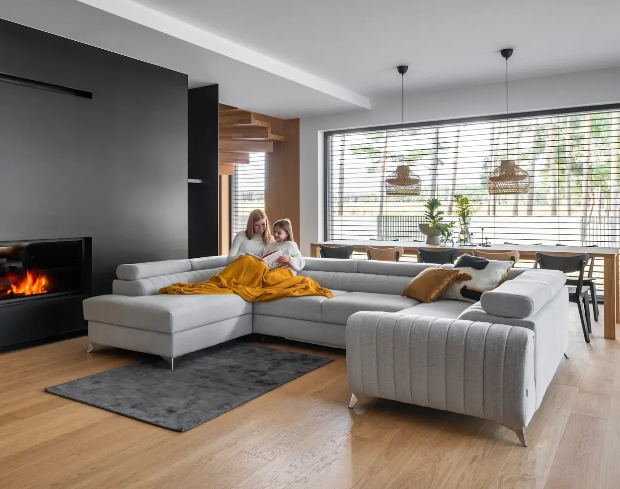 Jasnoszary narożnik Louis w kształcie litery U w nowoczesnym salonie z kominkiem. Na sofie mama z dzieckiem czytają książkę pod żółtym kocem, przy dużym oknie i jadalni.