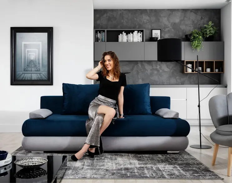 Kanapa rozkładana Milo w tkaninach Monolith 77 i Monolith 84 w kolorach niebieskim i szarym, ustawiona w nowoczesnym salonie. W tle widać zabudowę kuchenną, czarną lampę stojącą oraz dywan w szarej tonacji.