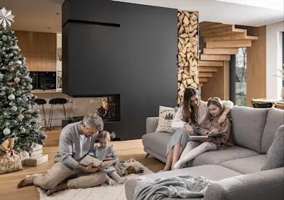 Rodzinne wnętrze w bożonarodzeniowym klimacie. Znajduje się w nim szary, rozkładany narożnik, na którym siedzą mama z córką. Obok, na dywanie, mężczyzna czyta książkę dziecku