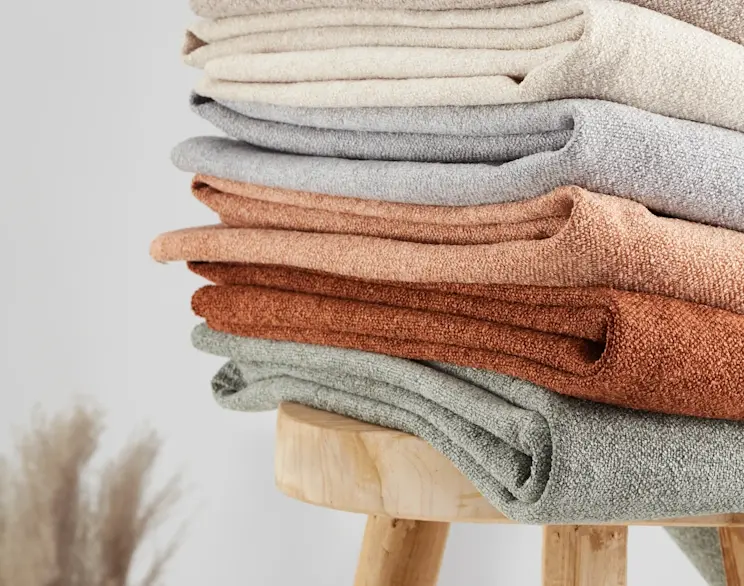 Tkanina boucle Xillar w różnych kolorach ułożona w stos na drewnianym stołku. Widoczne odcienie beżu, szarości i ciepłych brązów podkreślają miękką strukturę materiału, idealnego na obicia mebli.