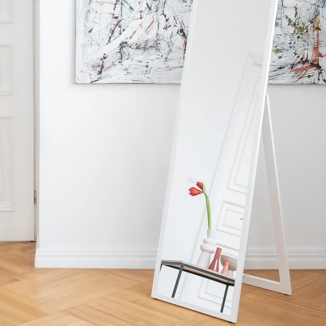 Stojące lustro Verona w białej ramie ustawione na podłodze w jasnym wnętrzu z drewnianym parkietem w jodełkę. W odbiciu widać wazon z tulipanem, obok stolik kawowy Moss i nowoczesne obrazy na ścianie.