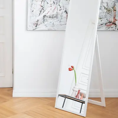 Stojące lustro Verona w białej ramie ustawione na podłodze w jasnym wnętrzu z drewnianym parkietem w jodełkę. W odbiciu widać wazon z tulipanem, obok stolik kawowy Moss i nowoczesne obrazy na ścianie.