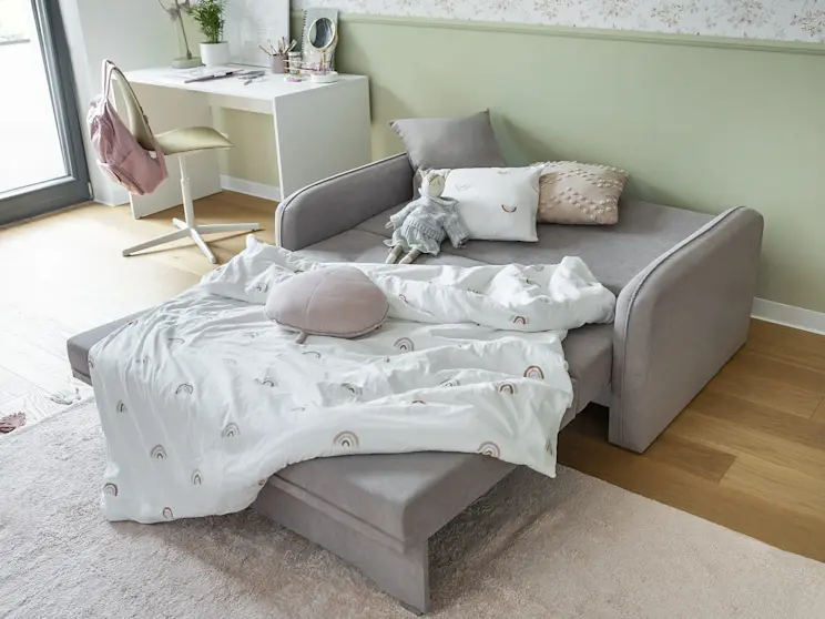 Pokój dziecka z rozkładaną kanapą Laine: szerokie siedzisko po rozłożeniu pełni rolę wygodnego łóżka. Jasna aranżacja z biurkiem Nevy, pastelową ścianą i dodatkami tworzy przytulne miejsce do odpoczynku.