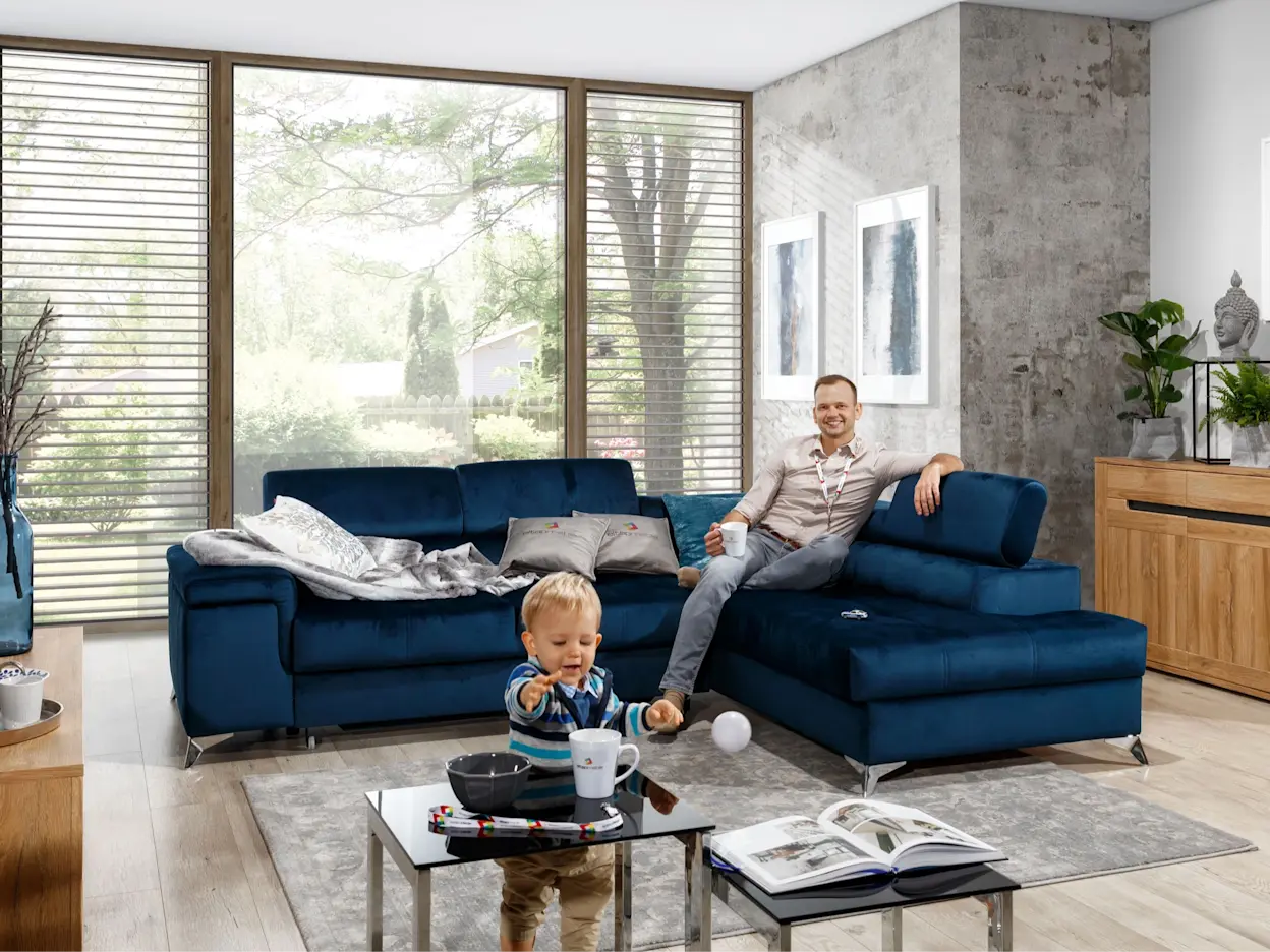 Duży narożnik Eridano w tkaninie Monolith 77 w jasnym, przestronnym salonie przy oknie; wygodna leżanka i wysokie oparcia sprawdzają się na drzemki, filmowe wieczory i rodzinny odpoczynek.